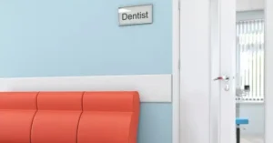 A blue-walled room with a sign reading Dentist above a door. An orange cushioned bench lines the wall. The door is slightly open, leading to a brighter area with blinds covering the windows.