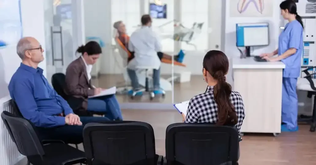 Three people are seated in a dental office waiting area, two taking notes. Through a glass partition, a dentist works with a patient in an exam chair. A dental assistant in scrubs uses a computer nearby.