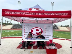 Two students sit at a table under a red Patterson Track and Field/Cross Country tent on a sports field. The tent showcases team values like teamwork, integrity, and accountability. Track stands and bleachers are visible in the background.