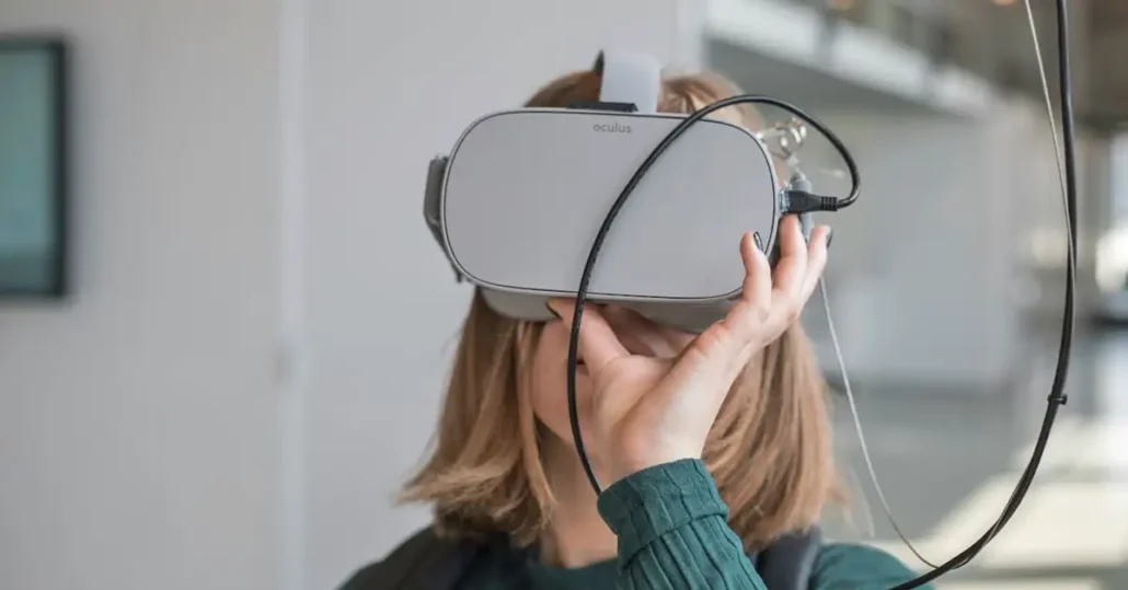 A person wearing a gray virtual reality headset, holding it with one hand. The background is blurred, showing an indoor setting with natural light coming from the right side. The individual is dressed in a dark green top.