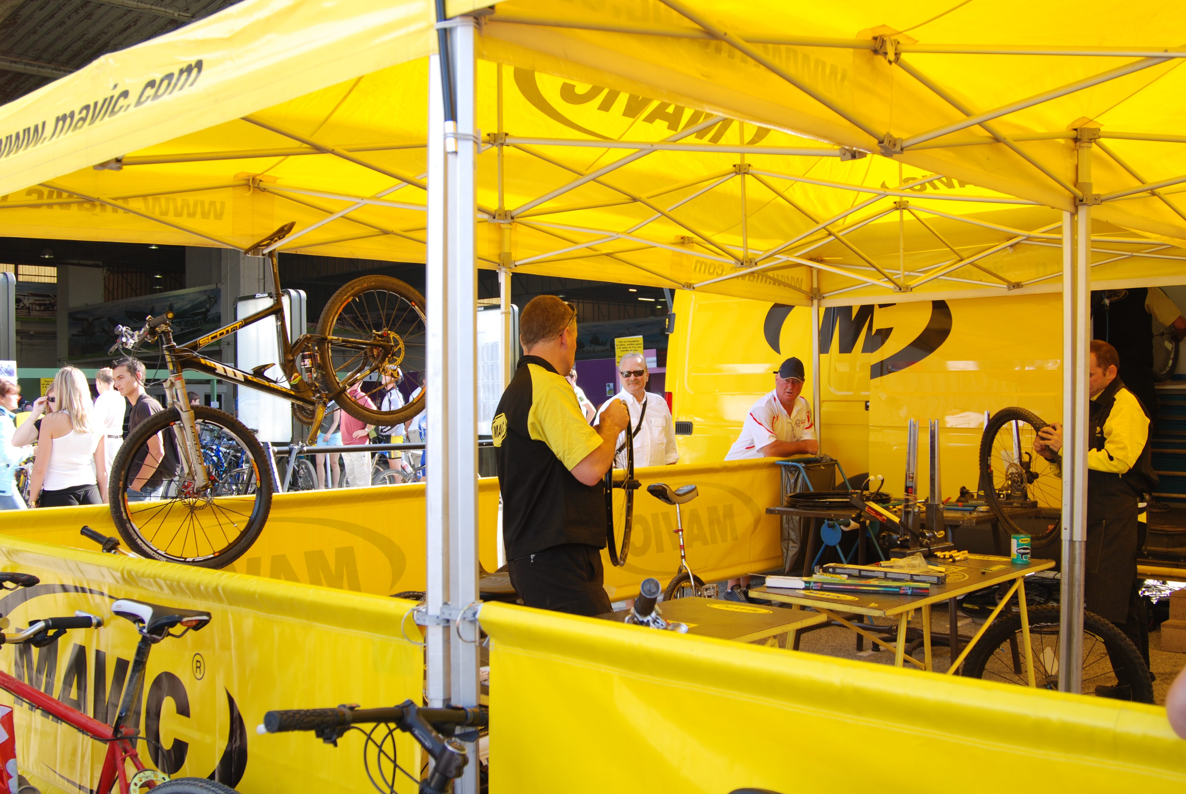 Mechanics in a yellow Mavic tent repair bicycles at an outdoor event, with tools and bike parts on tables. People and bikes can be seen in the background under sunny conditions.