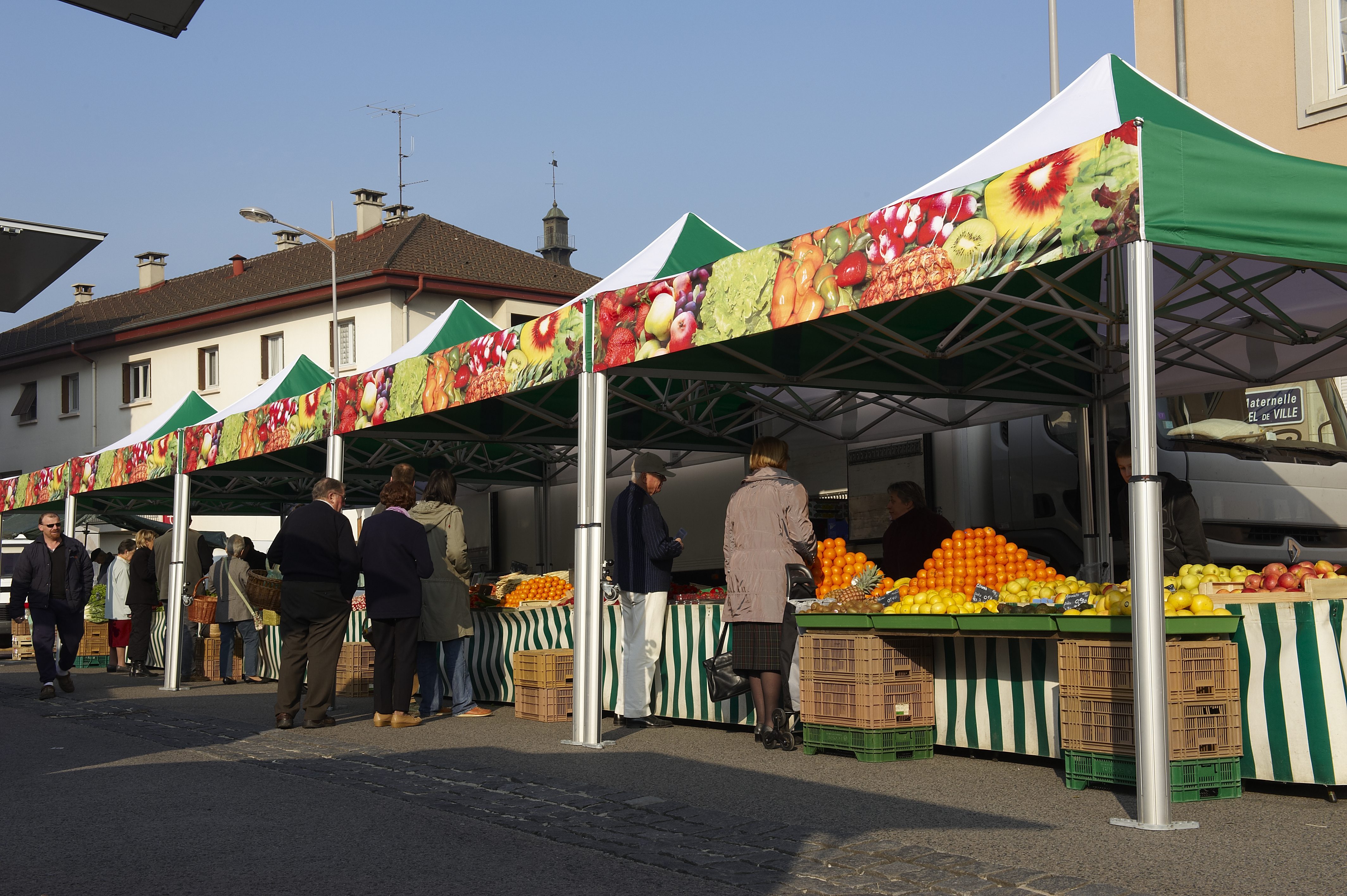 People shop for fresh produce at an outdoor market with striped stalls and colorful fruit decorations. Various fruits, including oranges and lemons, are displayed on tables. Buildings are visible in the background.