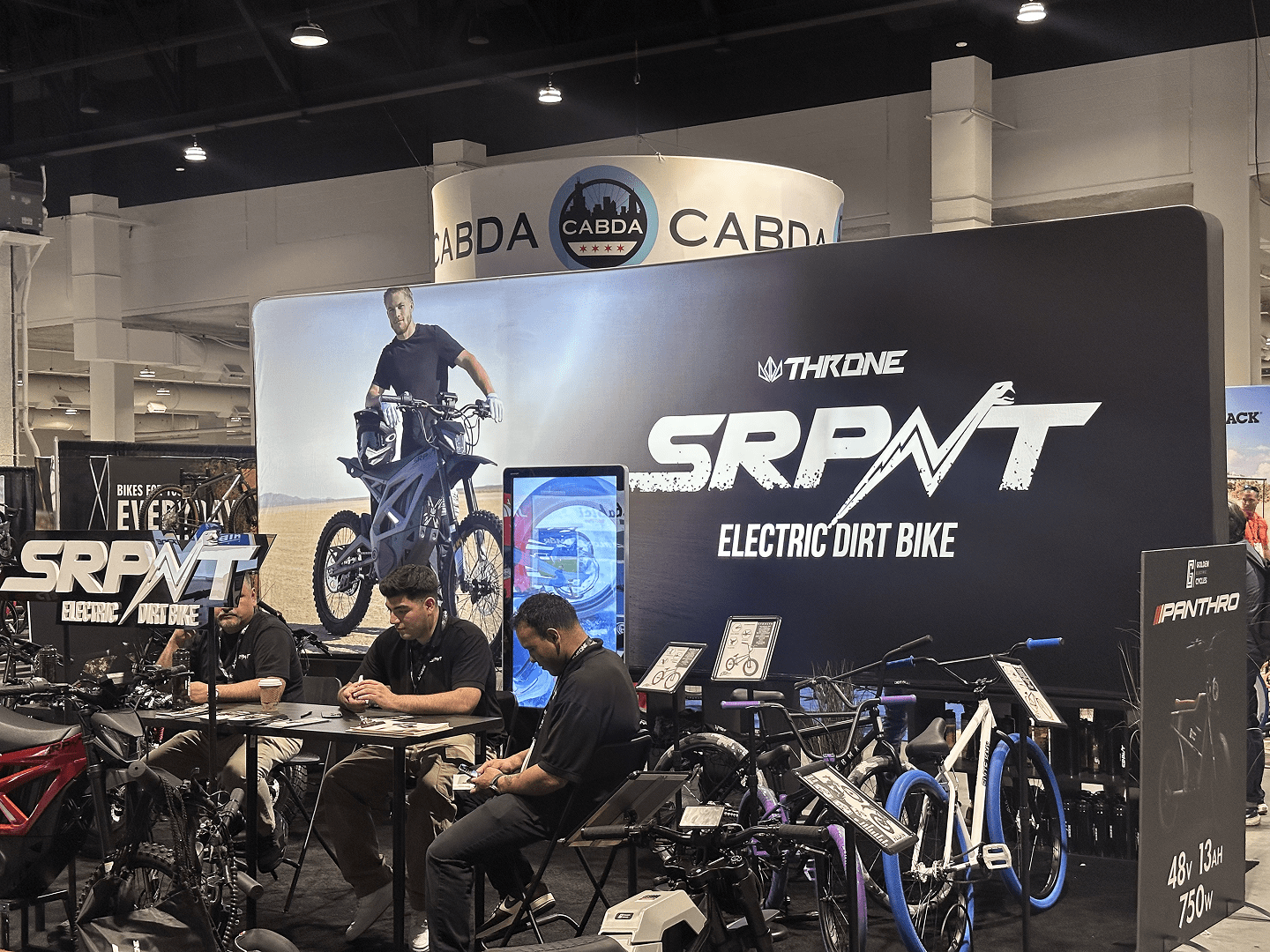 A booth at a convention displays electric dirt bikes and a large THRONE SRPNT banner. Three people sit at a table with bikes in front; signage advertises the SRPNT electric dirt bike. CABDA Expo branding is visible above.