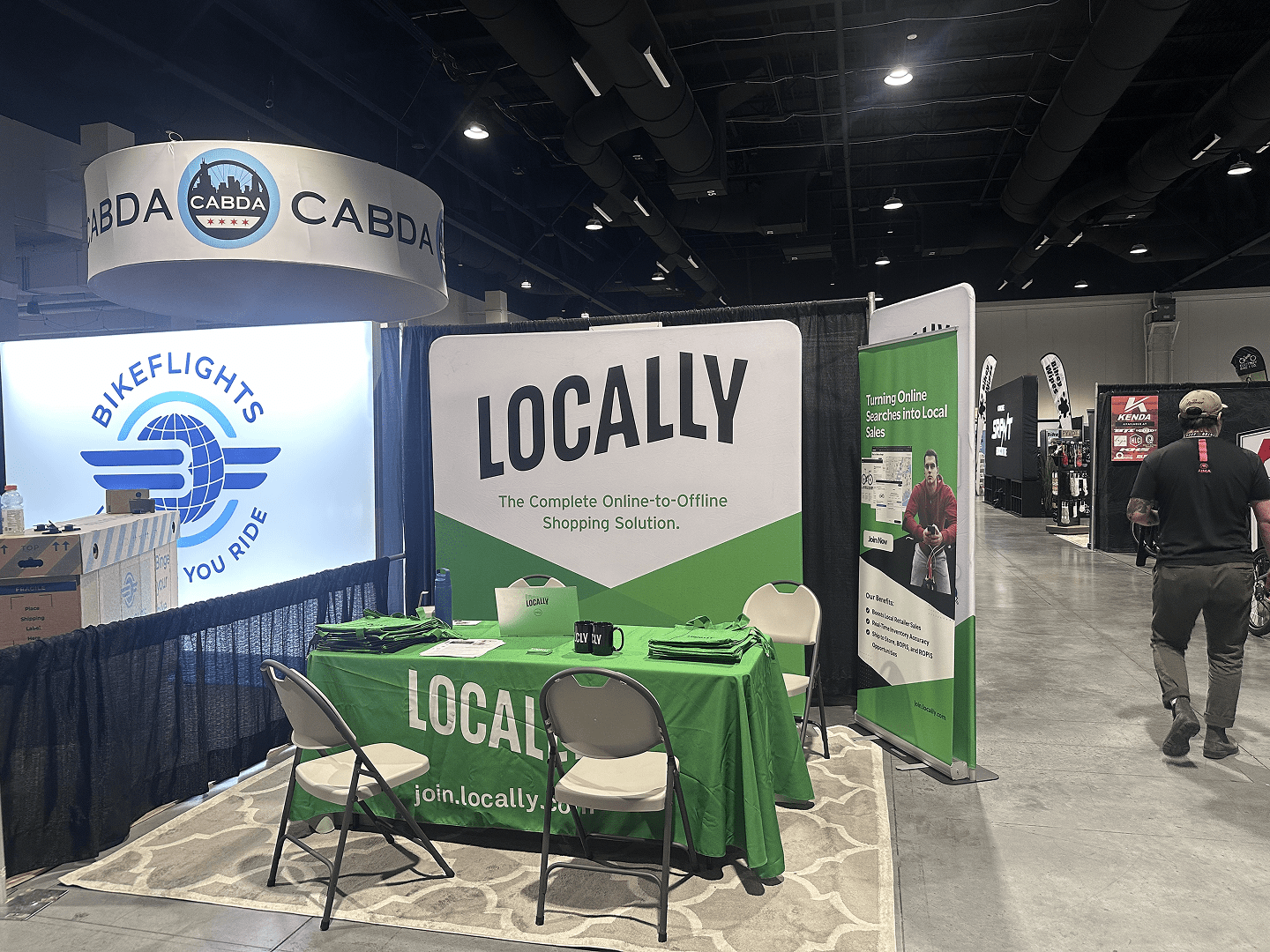 A trade show booth with a large LOCALLY sign and green tablecloth, informational pamphlets, and chairs. Nearby are other booths, including BIKEFLIGHTS and CABDA. The setting is an indoor exhibition hall.