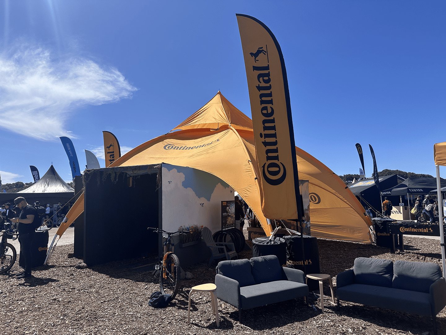 A large orange Continental-branded tent and flags at an outdoor event, with couches and bicycles in front, under a sunny blue sky. People and other booths are visible in the background.