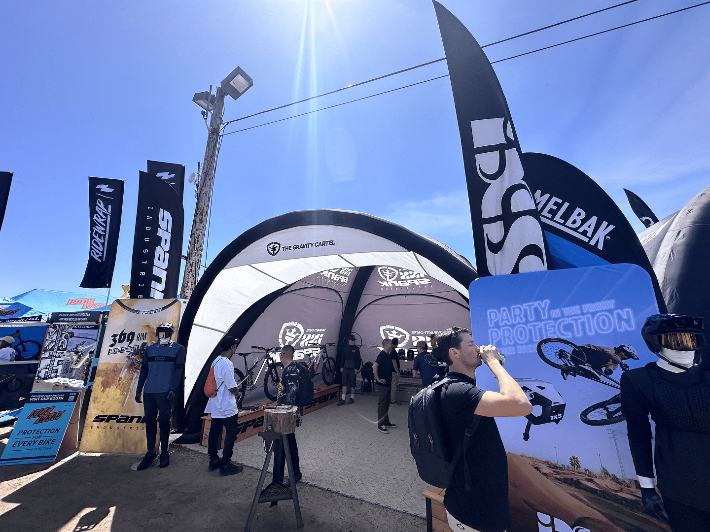 People gather outside and inside a branded event tent displaying bicycles and gear, with flags, banners, and stands advertising bike products under a clear blue sky. A mannequin in a helmet stands near a sign reading PARTY—PROTECTION.