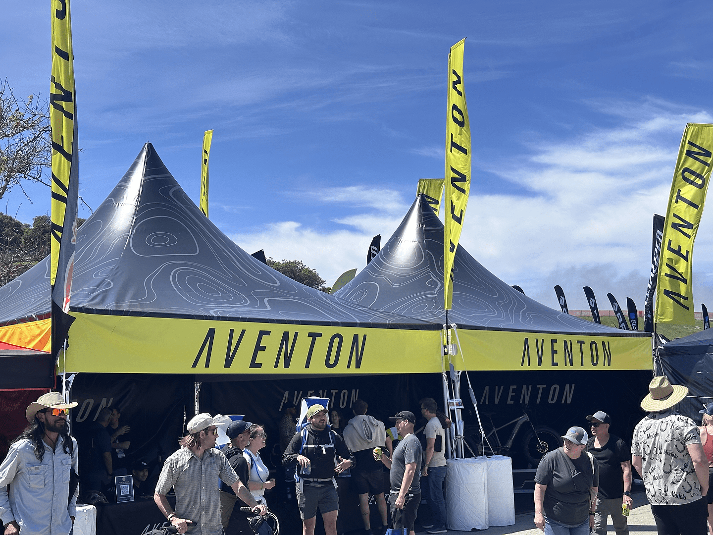 A crowd of people walk by a large outdoor tent with black and yellow AVENTON banners and flags at an event under a blue sky.