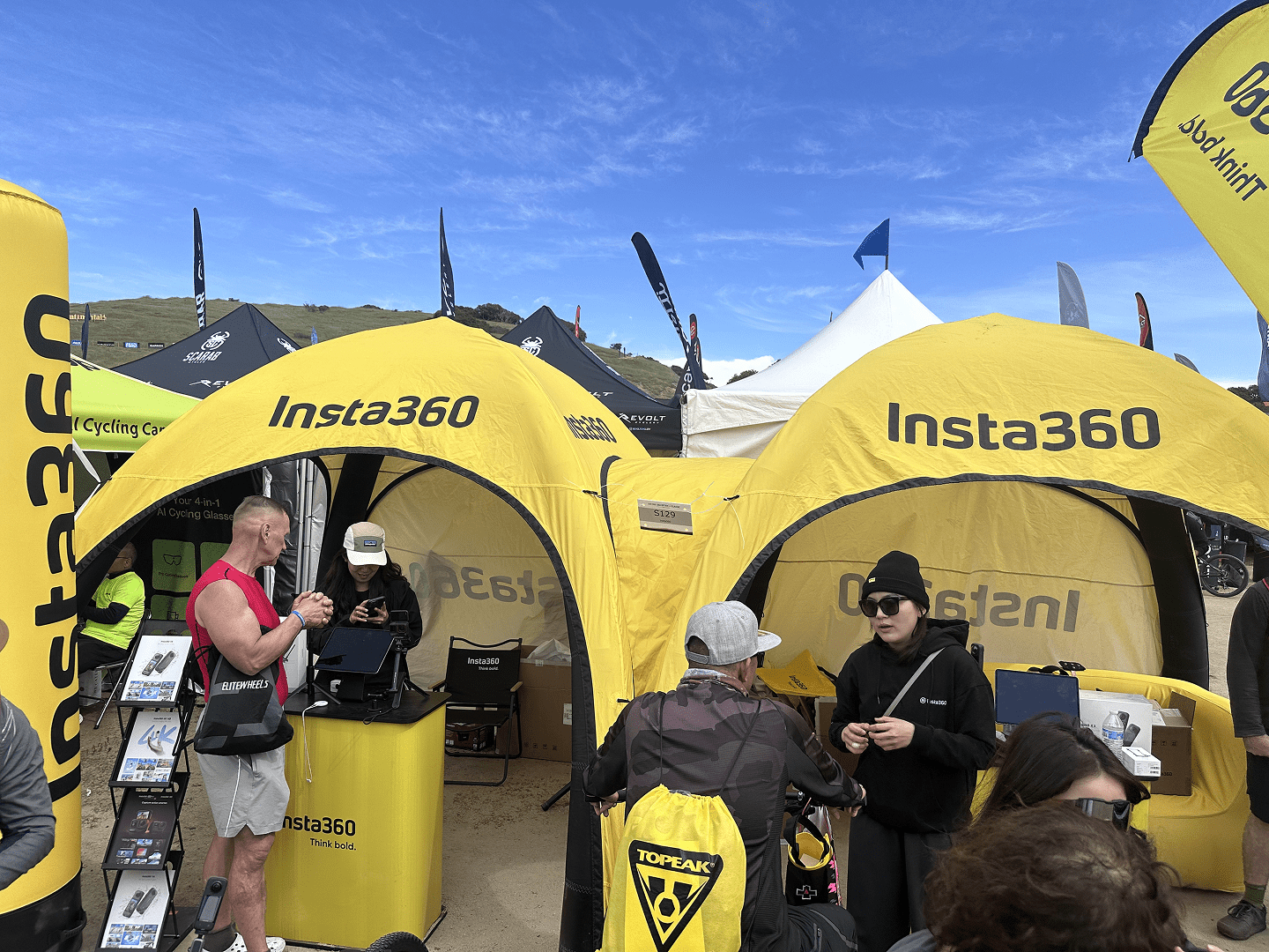 People gather around two bright yellow Insta360 tents at an outdoor event. Some interact with products, while others stand and observe under a clear blue sky with hills in the background.