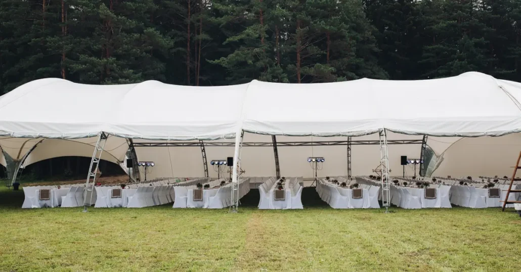 A large white event tent with long banquet tables and chairs set up underneath, situated on a grassy area with a forest of tall trees in the background.