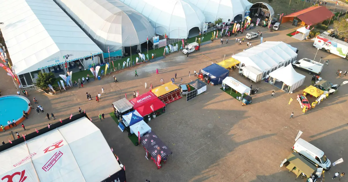 Aerial view of an outdoor event with large white tents, vendor booths, people walking, and a circular pool on the left. Vehicles and banners are visible throughout the open, sunlit area.