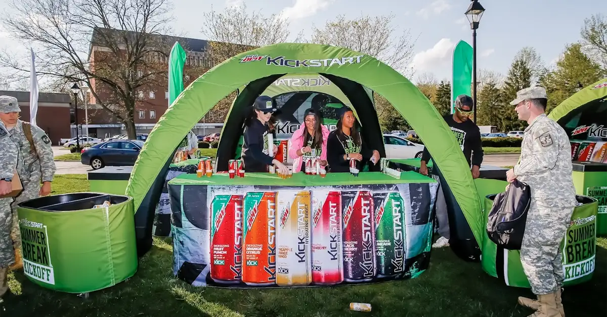 People in military uniforms and others gather at a green outdoor tent promoting Mountain Dew Kickstart energy drinks, with bottles displayed on the table and banners in the background.