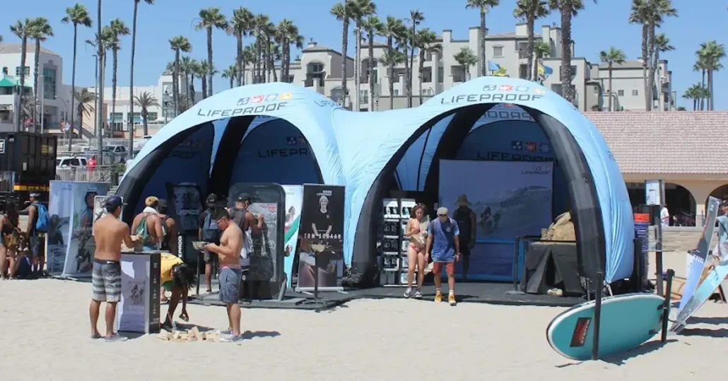 People gather at a beach event with two large blue LifeProof Air Dome tents, palm trees, beach equipment, and nearby buildings in the background. Some people are setting up games and surfboards are visible.