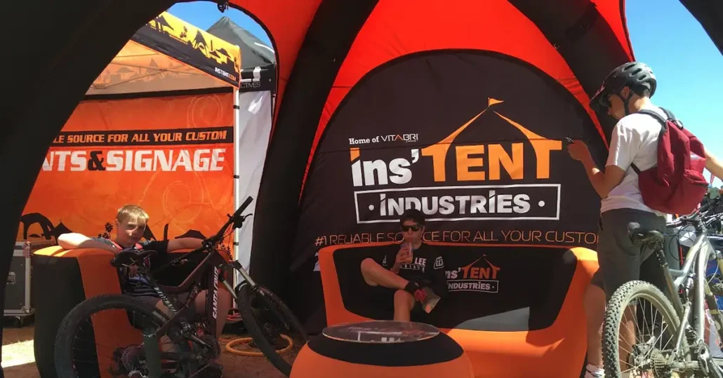 Two kids sit relaxing under a branded tent with Instent Industries signage, surrounded by bikes. One child uses a phone, while the other sits with sunglasses and a bike, in a sunny outdoor event area.