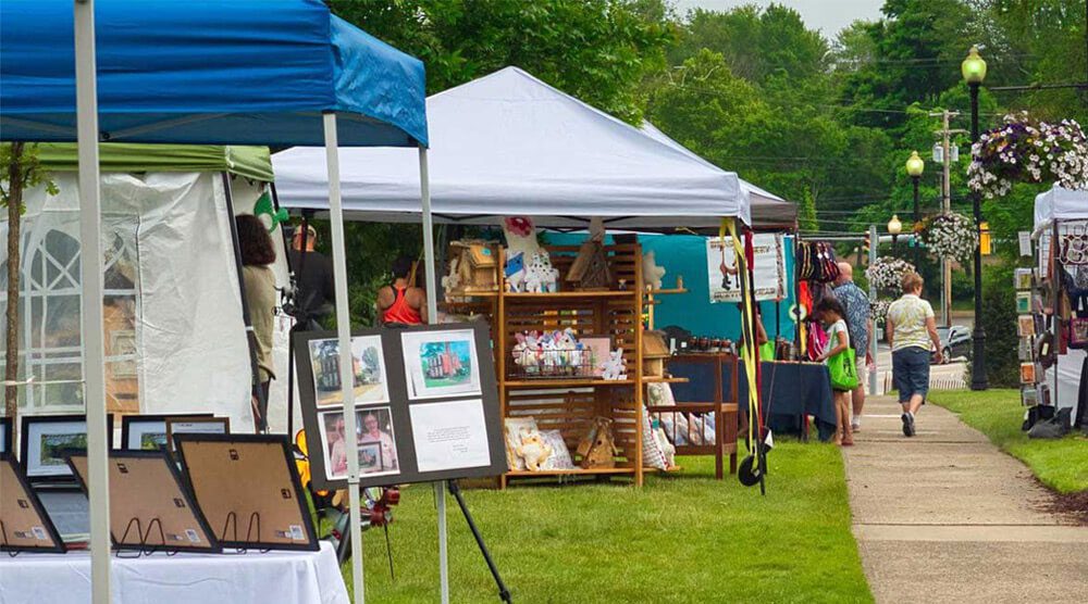 Outdoor art fair with tents displaying framed artwork, crafts, and handmade items along a grassy sidewalk. People walk by, browsing the booths on a cloudy day with lush green trees in the background.