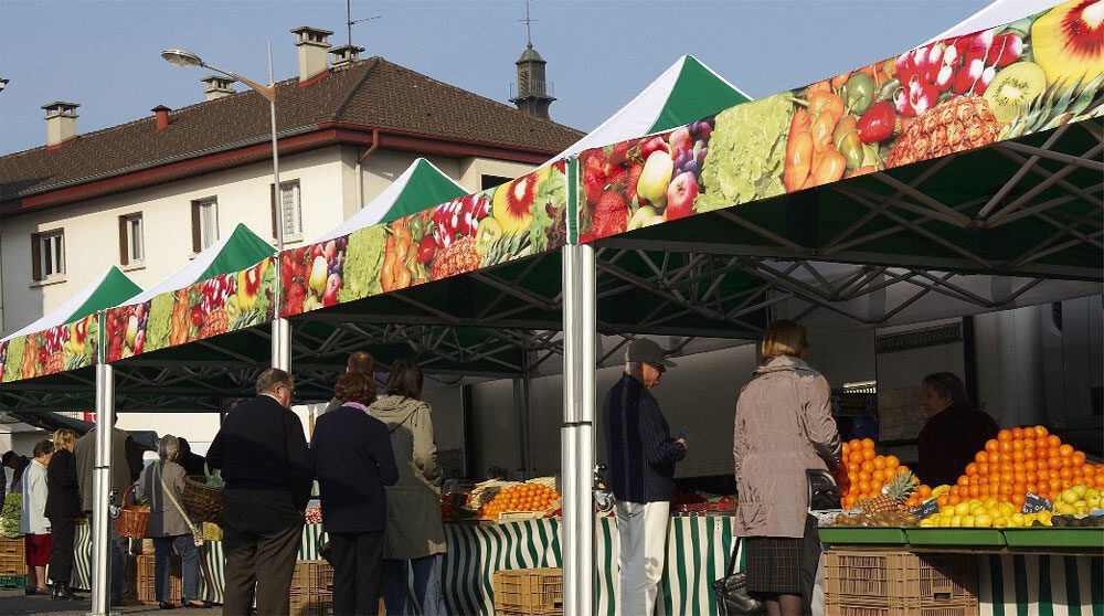 People shop for fresh produce at an outdoor market with stalls decorated with fruit and vegetable images. Oranges and other fruits are displayed on tables under striped canopies. Buildings are visible in the background.