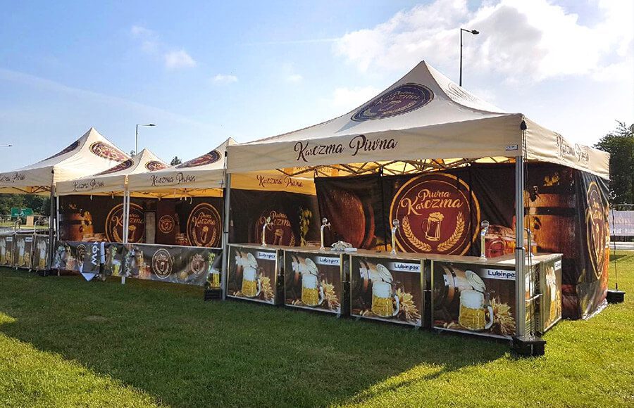 Outdoor event tents with Kazma Pivna branding, displaying beer graphics and signage. Counters are set up for serving drinks, and the tents are on a grassy area under a partly cloudy sky.