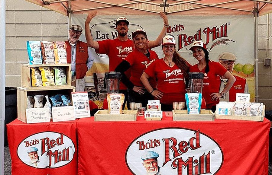 Six smiling people in red Bob’s Red Mill shirts stand behind a booth displaying various Bob’s Red Mill products under a branded canopy at an outdoor event. The booth has products like protein powders and cereals.