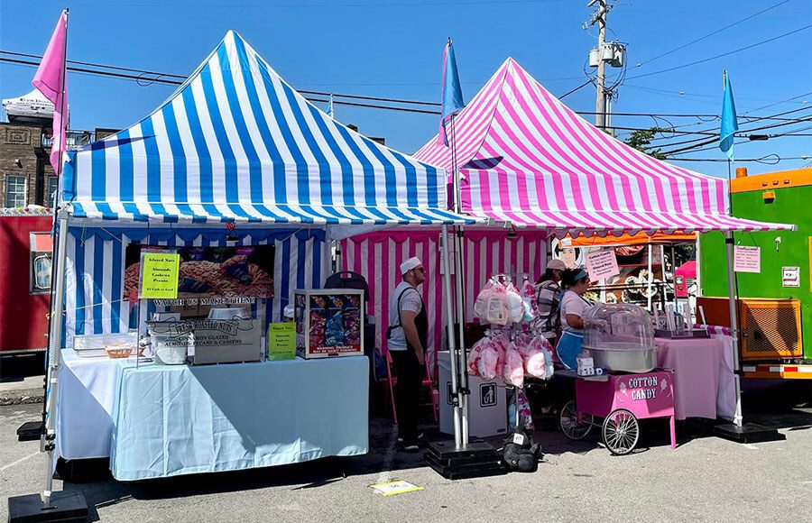 Two outdoor market stalls with blue-and-white and pink-and-white striped tents. Vendors sell baked goods and cotton candy, with displays and machines visible. The scene is sunny with clear skies.