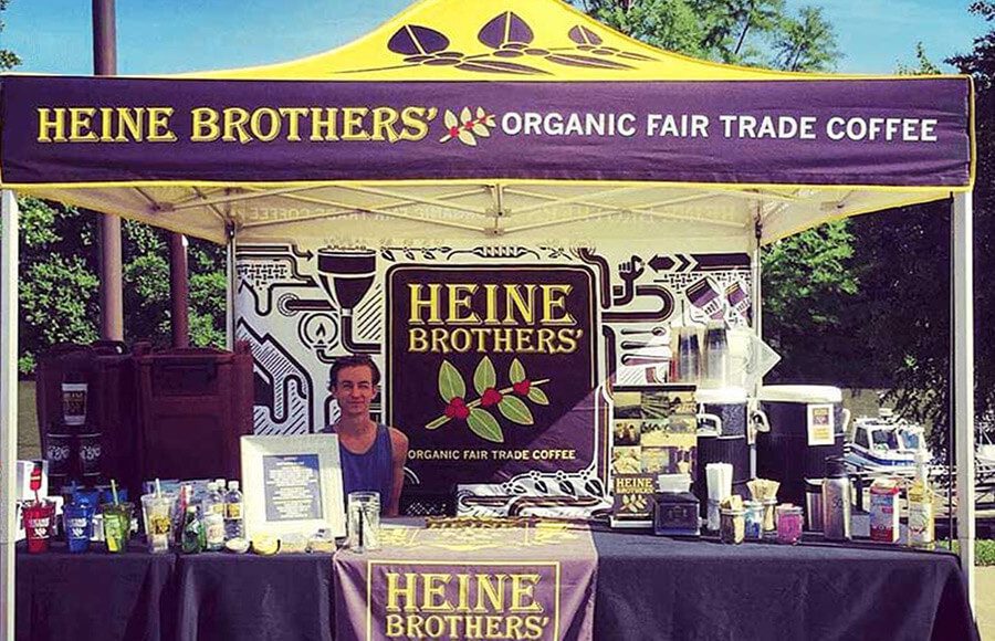 A person stands behind a booth under a yellow and purple tent branded Heine Brothers Organic Fair Trade Coffee, with coffee products and promotional materials displayed on the counter.