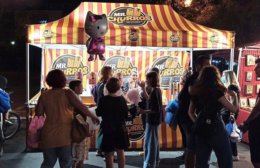 People gather at a brightly lit Mr. Churros stand with a yellow and red striped canopy at night. A Hello Kitty balloon and cotton candy are visible among the crowd. The atmosphere is lively and festive.