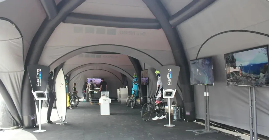 Large event tent with arched ceilings, GoPro display stations, screens showing action footage, and cyclists in helmets and gear interacting with staff and bikes. The space appears set up for a cycling or tech event.