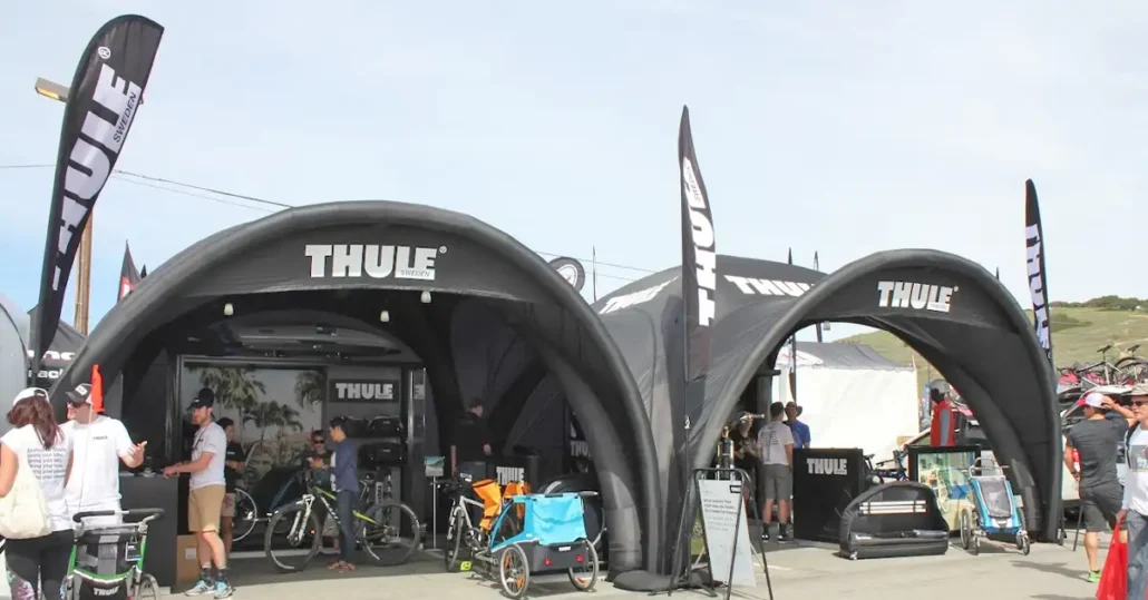 Two black inflatable Thule AirDome tents at an outdoor event display bike racks, strollers, and accessories. People are gathered around, some talking and browsing products, with Thule flags and banners visible.