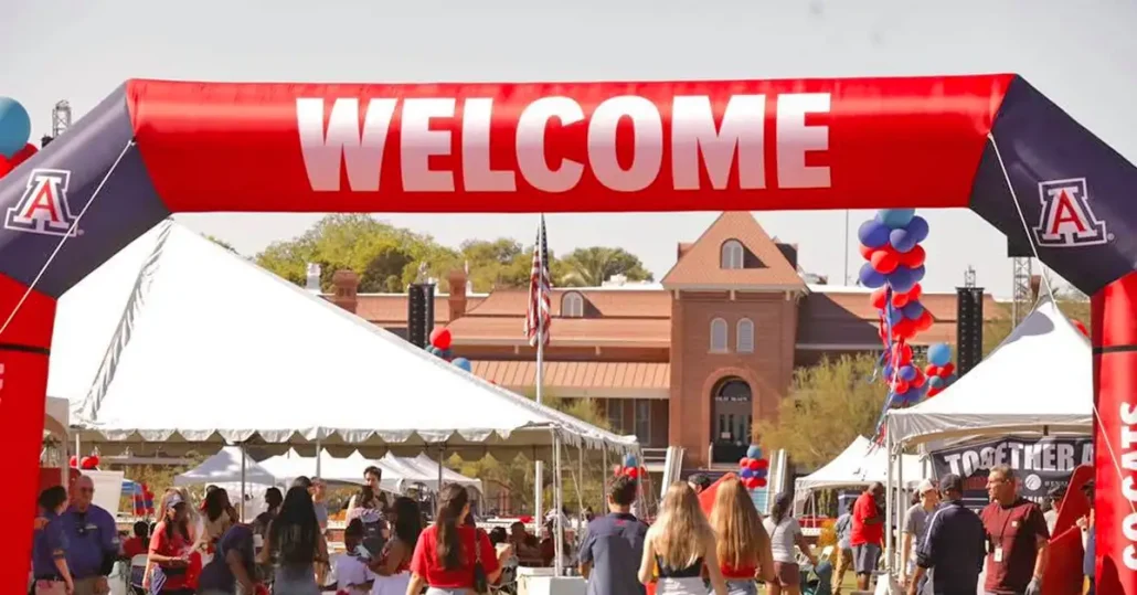 Students walk under a large red “WELCOME” inflatable arch at an outdoor event on the University of Arizona campus, with tents, balloons, and the campus building in the background.