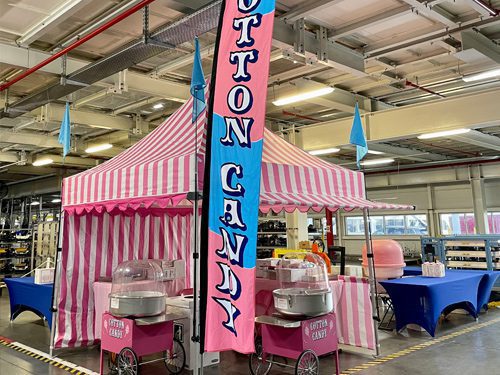 A pink and white striped tent with a blue and pink Cotton Candy banner stands inside a large indoor space, flanked by cotton candy machines and tables covered with blue cloths.