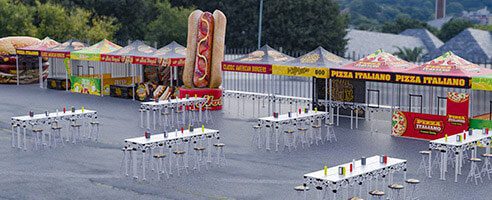 Outdoor food court with colorful booths offering pizza and hot dogs, including a large hot dog statue, and several rows of white tables with attached stools and condiments on top. Trees and rooftops are visible in the background.
