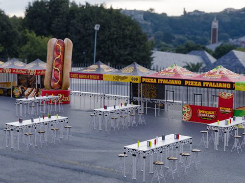 Several empty outdoor tables and stools are set up in front of colorful food stalls, including a large hot dog display and pizza stands, at what appears to be a food festival or market. Trees and buildings are in the background.