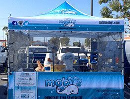 A blue and white food stand tent labeled Moby’s Original Fish Sandwich with a whale logo, set up outdoors. People are working inside the tent, and cars are parked in the background.