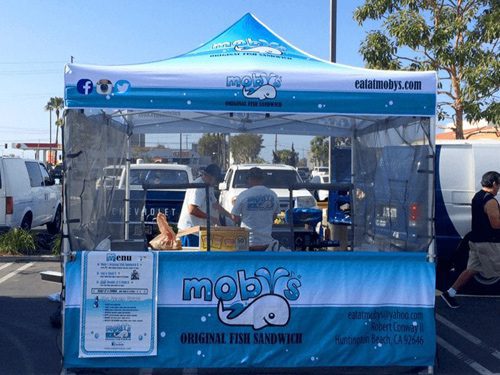 A blue and white tent labeled mobys ORIGINAL FISH SANDWICH serves food in a parking lot. Staff prepare food inside. The tent has social media icons and contact info. Cars and palm trees are visible in the background.