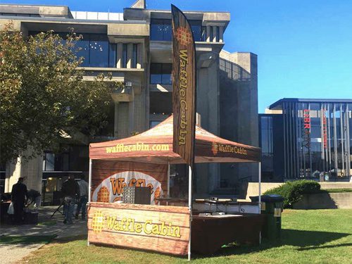 A Waffle Cabin food stand with a branded canopy and flag is set up outdoors on a sunny day, in front of a modern concrete building. The stand appears ready for customers.