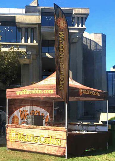 A pop-up tent labeled “Waffle Cabin” displays a waffle pattern and logo on a college campus lawn, with a tall vertical flag and waffle toppings visible on the counter.