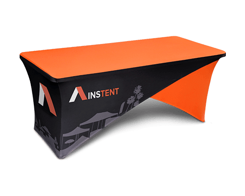 A rectangular table covered with a fitted, branded tablecloth featuring the AINSTENT logo. The tablecloth is mostly black with orange accents and a graphic of tents; the tabletop is solid orange.