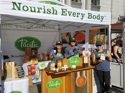 Three people stand smiling at a Pacific Foods booth, holding product samples. The booth canopy reads “Nourish Every Body,” and a table displays various Pacific Foods packaged products.
