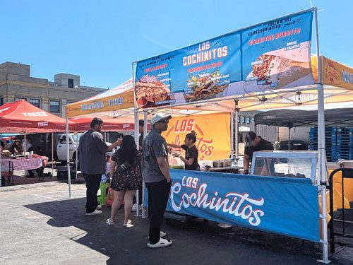 A group of people order food at the Los Cochinitos stand under blue and orange tents at an outdoor market on a sunny day. A large sign displays menu options above the counter.
