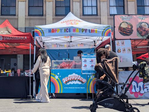 A colorful Happy Ice Happy Life dessert stand sells frozen treats at an outdoor market. People, including a woman in beige and a person pushing a stroller, stand nearby in front of a building with food signs.