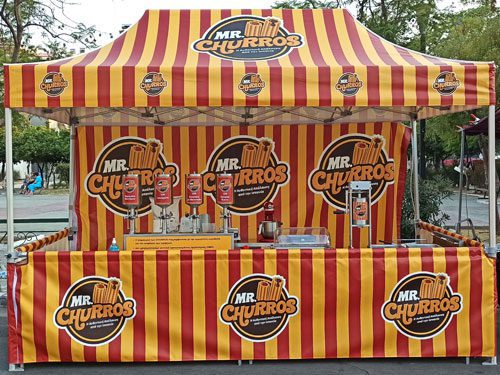 A striped red and yellow Mr. Churros booth with churro-making machines and dispensers on the counter, set outdoors with trees in the background. The booth is decorated with large Mr. Churros logos.