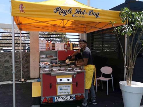 A man wearing a yellow apron prepares food at a red and yellow Rayli Hot-Dog stand under a bright yellow canopy, with condiments and a potted plant nearby.