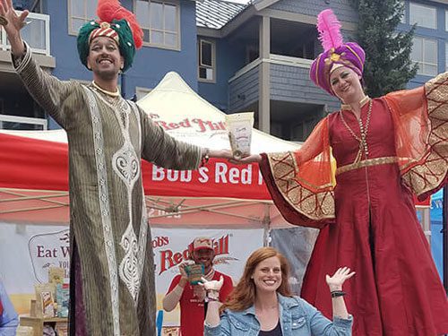 Two people in colorful, ornate costumes and tall hats pose on stilts in front of a Bob’s Red Mill booth, with a smiling woman kneeling between them and another person standing in the background.