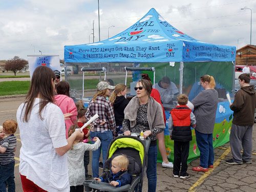 A group of people, including adults and children, gather around a colorful tent labeled DEAL OF THE DAY in a parking lot. Some adults talk while kids stand nearby; a woman pushes a stroller in the foreground.