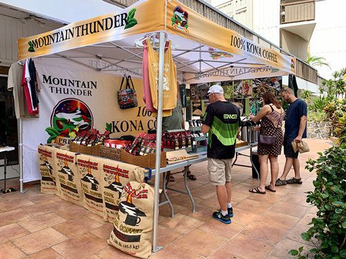 A coffee stand with a yellow Mountain Thunder 100% Kona Coffee canopy displays bags, shirts, and coffee products. Four people browse the items outside in a sunny, outdoor market setting.