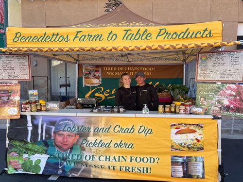 A yellow farmers market stall labeled “Benedetto’s Farm to Table Products.” Two people stand behind a counter displaying jars and produce. Signs advertise lobster and crab dip, pickled okra, and “We Make It Fresh!”.