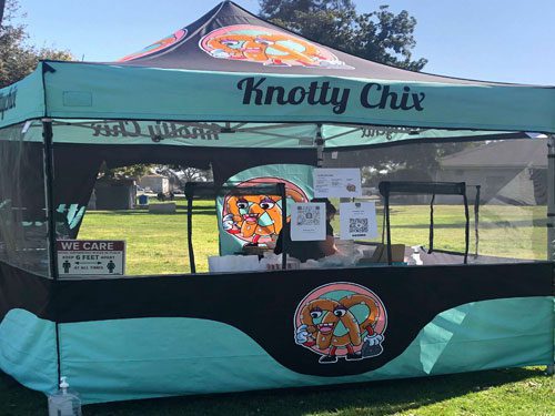 A teal and black tent labeled Knotty Chix at an outdoor event, with a cartoon pretzel mascot on the canopy. Signs and food items are visible inside the booth, and a “6 feet” social distancing sign is displayed.