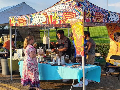 A woman in a floral dress speaks with a vendor at a colorful outdoor market booth under a tent labeled Yazoo Yaupon. The table displays jars, bottles, and baskets, and other tents are visible in the background.