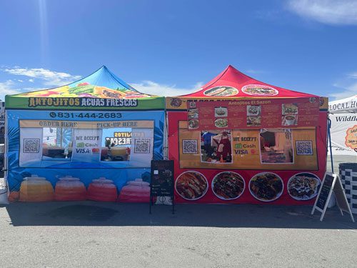 Two colorful outdoor vendor tents selling food and drinks, decorated with images of aguas frescas and various Mexican dishes. Signs display phone numbers and accept cash and Visa payments. The sky is clear and sunny.