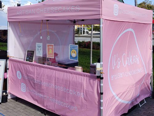 A pink market stall with Vs Cakes Artisan Cheesecakes on the canopy and tablecloth. Cheesecakes and signs are displayed on the table inside the booth at an outdoor event.