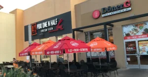Outdoor seating area with black tables and chairs, shaded by red and orange umbrellas, in front of two restaurants: Poke One N Half (Fresh Fish) and Döner G (Turkish & Mediterranean Grill).