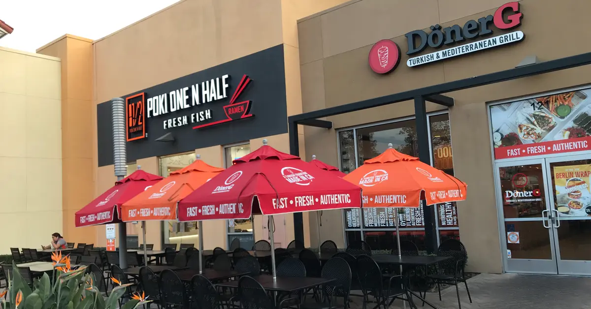 Outdoor seating area with black tables and chairs, shaded by red and orange umbrellas, in front of two restaurants: Poke One N Half (Fresh Fish) and Döner G (Turkish & Mediterranean Grill).